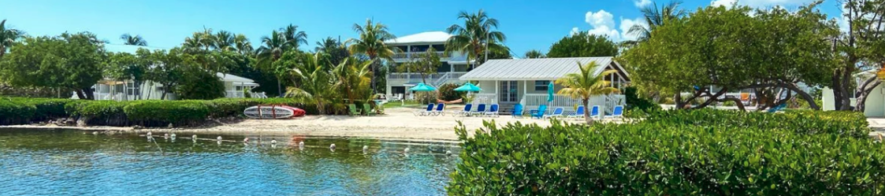 A serene beach scene with clear water, lush greenery, and houses in the background under a bright blue sky with fluffy clouds.