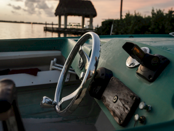 A boat's steering wheel and controls against a sunset over water, with a tiki hut and greenery in the background.