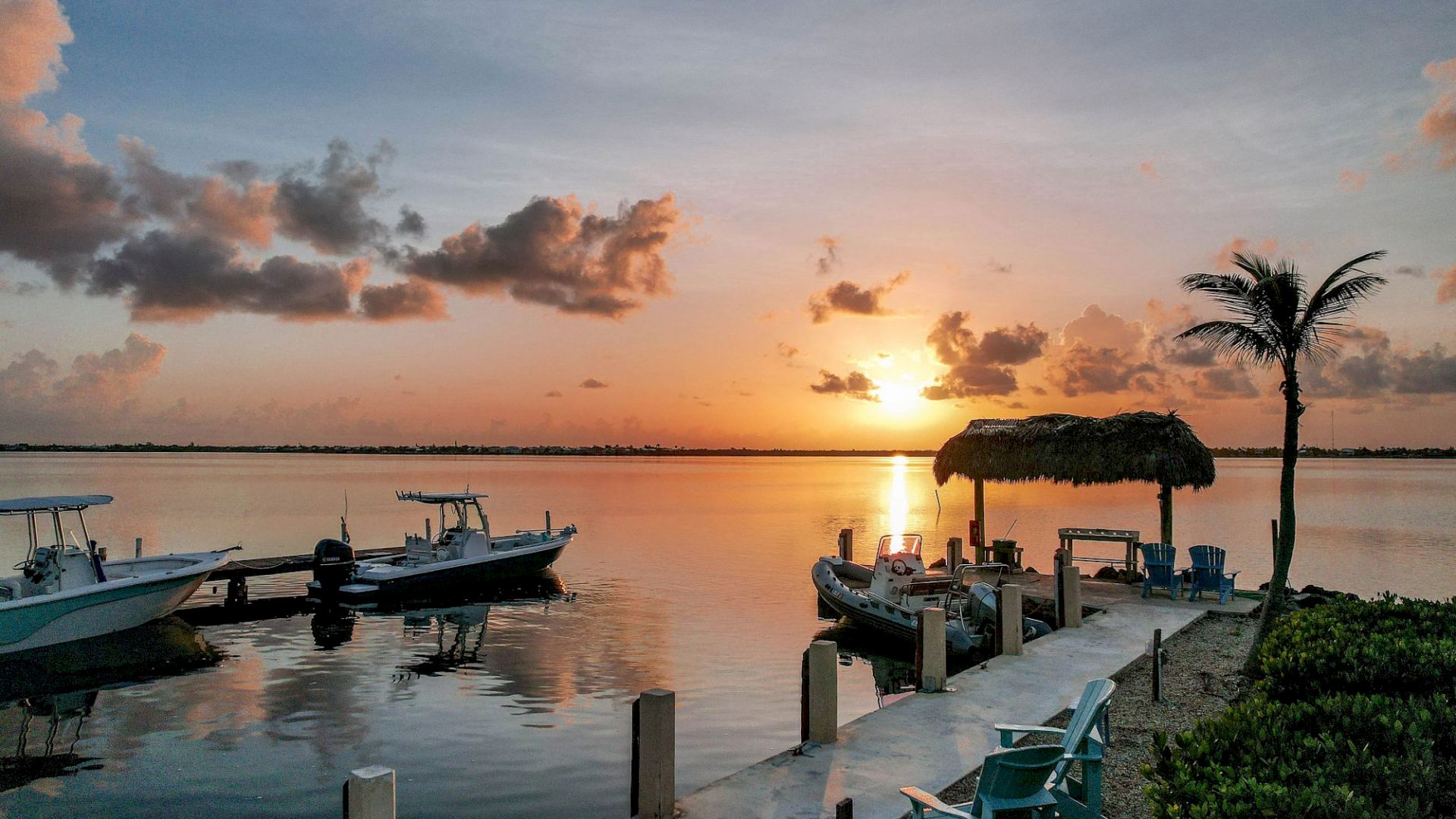Sunset over a calm bay with docked boats, a palm tree, and a thatched hut on the pier.