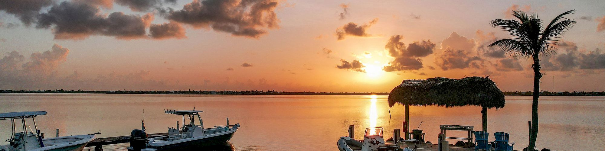 Sunset over a calm bay with docked boats, a palm tree, and a thatched hut on the pier.