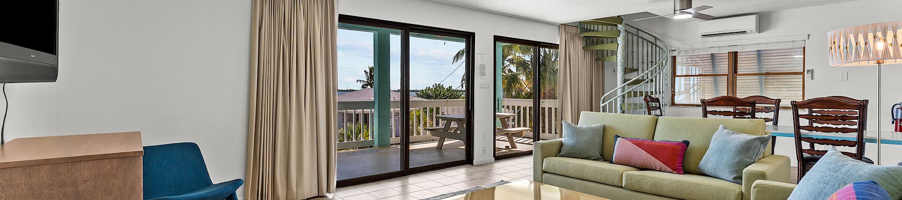 Bright living room with modern furniture, colorful sofas, a wooden coffee table, ceiling fan, and large glass doors opening to a deck.
