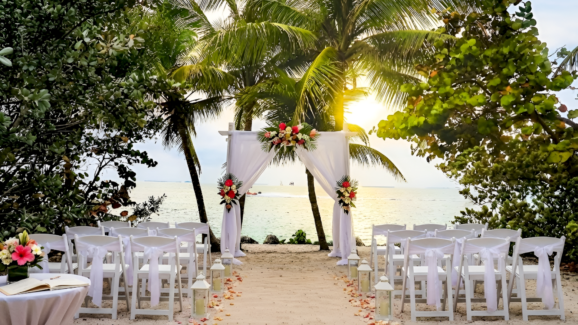 A beach wedding setup with white chairs, a floral arch, and sunset light over the ocean, on a sandy aisle.