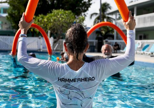Person wearing a white shirt with &ldquo;HAMMERHEAD&rdquo; on back, standing in a pool and holding up red pool noodles like a hoop, sunny day.