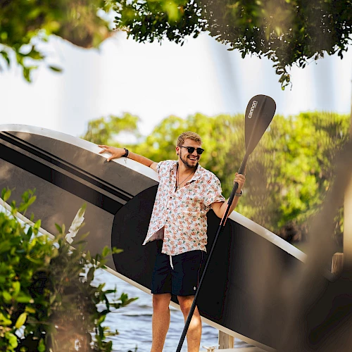 A man carrying a large stand-up paddleboard and paddle, walking outdoors among trees, wearing a patterned shirt and shorts.