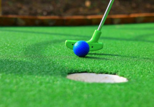 A blue golf ball near a green putter on a bright green putting surface, with a hole in the foreground.