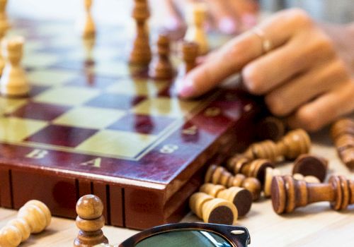 A person plays chess on a wooden board with scattered pieces, while sunglasses and a ring rest on the table nearby.