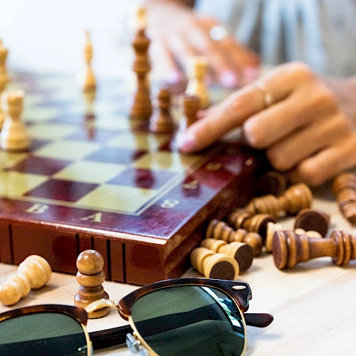 A person plays chess on a wooden board with scattered pieces, while sunglasses and a ring rest on the table nearby.