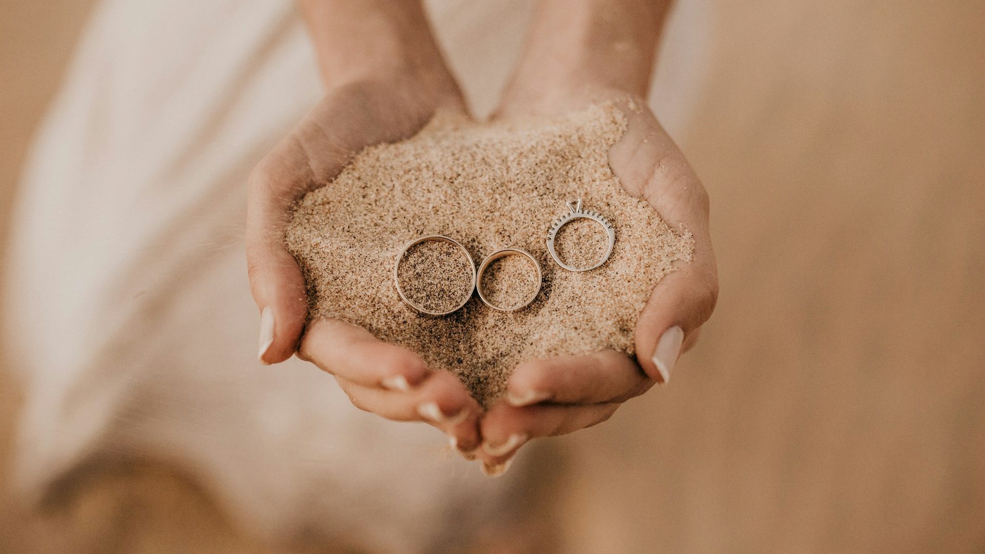 Two hands hold a mound of sand with three rings nestled in it.