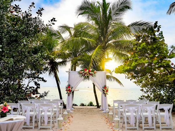 A beach wedding setup with white chairs and a flower-decorated arch, sandy aisle lined with petals, palm trees, and the sun setting over the sea.