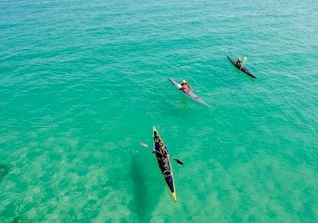 Three surfers/paddlers float on clear turquoise water, one on a yellow board, others on dark boards, aerial view.