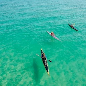 Three surfers/paddlers float on clear turquoise water, one on a yellow board, others on dark boards, aerial view.