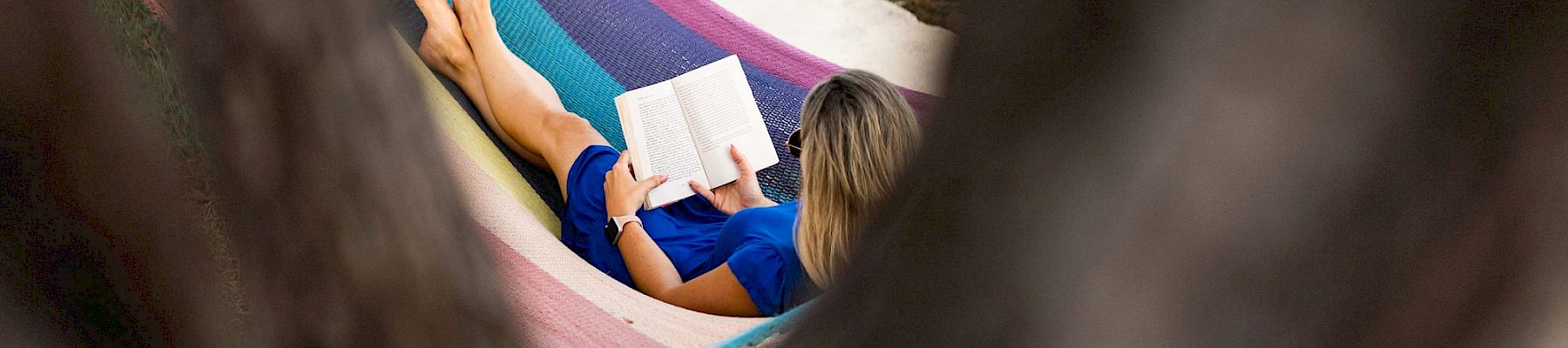 A person relaxing in a striped hammock by the beach, reading a book, framed by tree branches in the foreground.