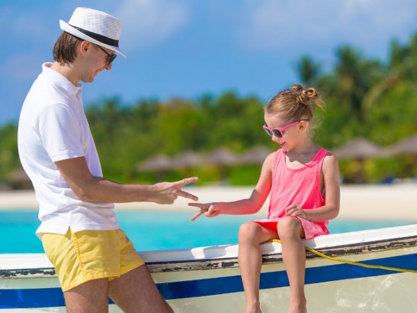 Two kids on a boat by a sunny beach share a high-five; girl in pink sits, boy in white tee and yellow shorts, tropical backdrop.