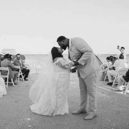 A black-and-white wedding on the beach: a bride and groom kiss as guests sit in rows along the shore, celebrating beneath the open sky.