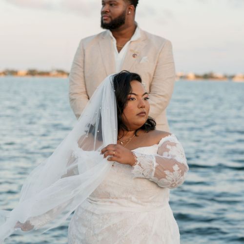 A couple dressed in wedding attire by the water: the bride in a lace white dress and veil, the groom in a light suit, standing by a lake or sea.