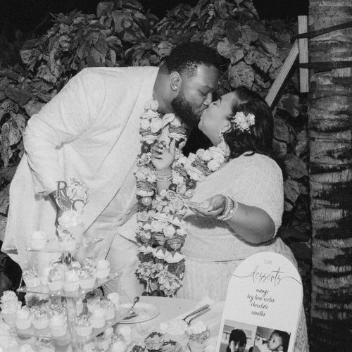 A couple kisses at a wedding cake table, wearing leis and a floral backdrop. They share a sweet moment amid cupcakes and a photo frame.
