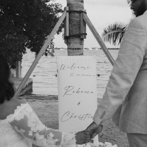 A couple in light attire walk hand-in-hand toward a sign with names, outdoors by the water and a palm tree, wedding vibe.