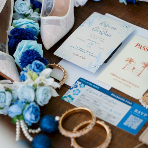 A bridal scene featuring white and blue flowers, a pair of white shoes, wedding invitations, blue cards, and some jewelry on a wooden surface.