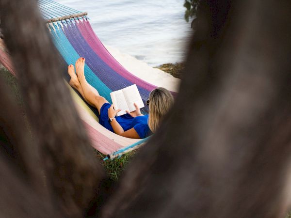 Someone relaxing in a colorful striped hammock by the water, reading a notebook, framed by tree branches.