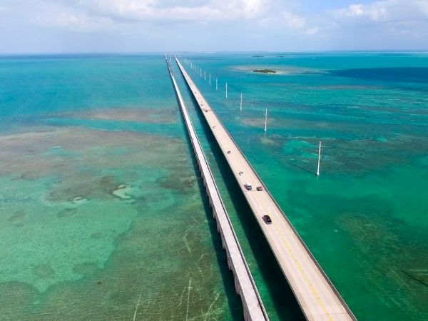 A long bridge splits turquoise water, connecting land over a shallow reef, with utility poles marching into the horizon under a sunny sky.