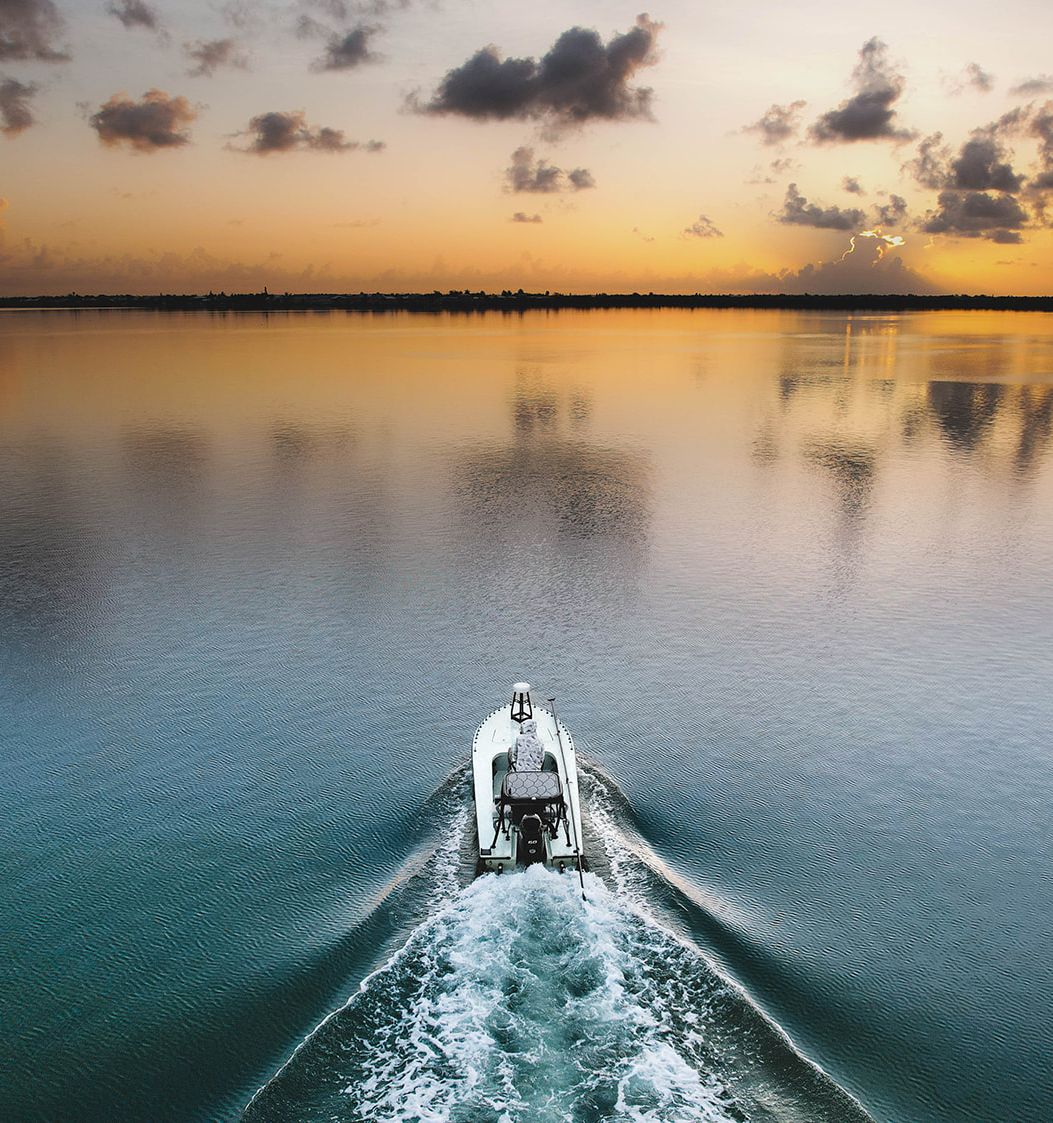 A small boat glides across calm water at sunset, leaving a frothy wake as the horizon glows with orange and pink clouds.