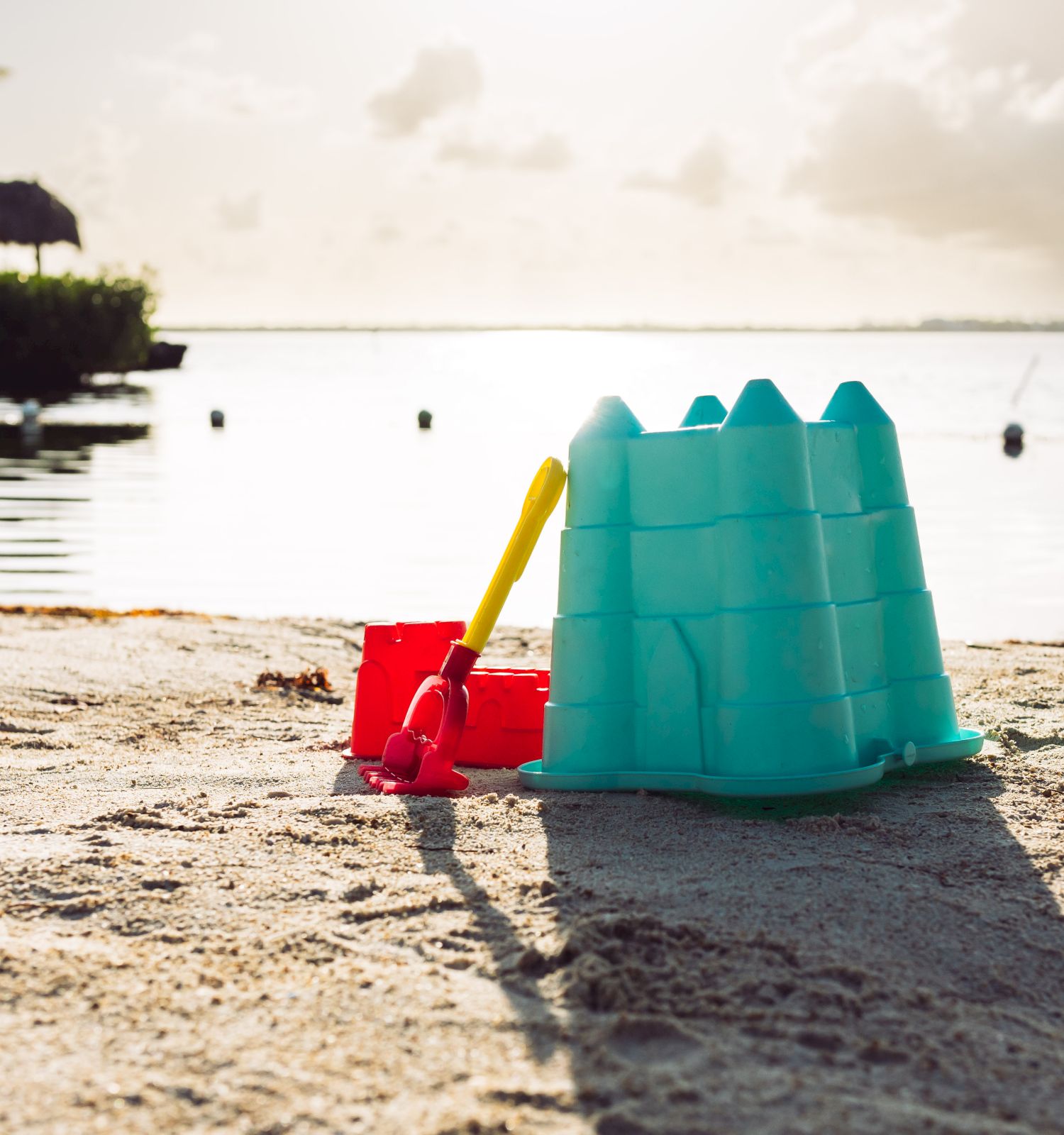 A bright teal sandcastle, a red bucket, and a yellow shovel on a sunny beach with calm water and a distant palm hut at sunset, ending with a period.