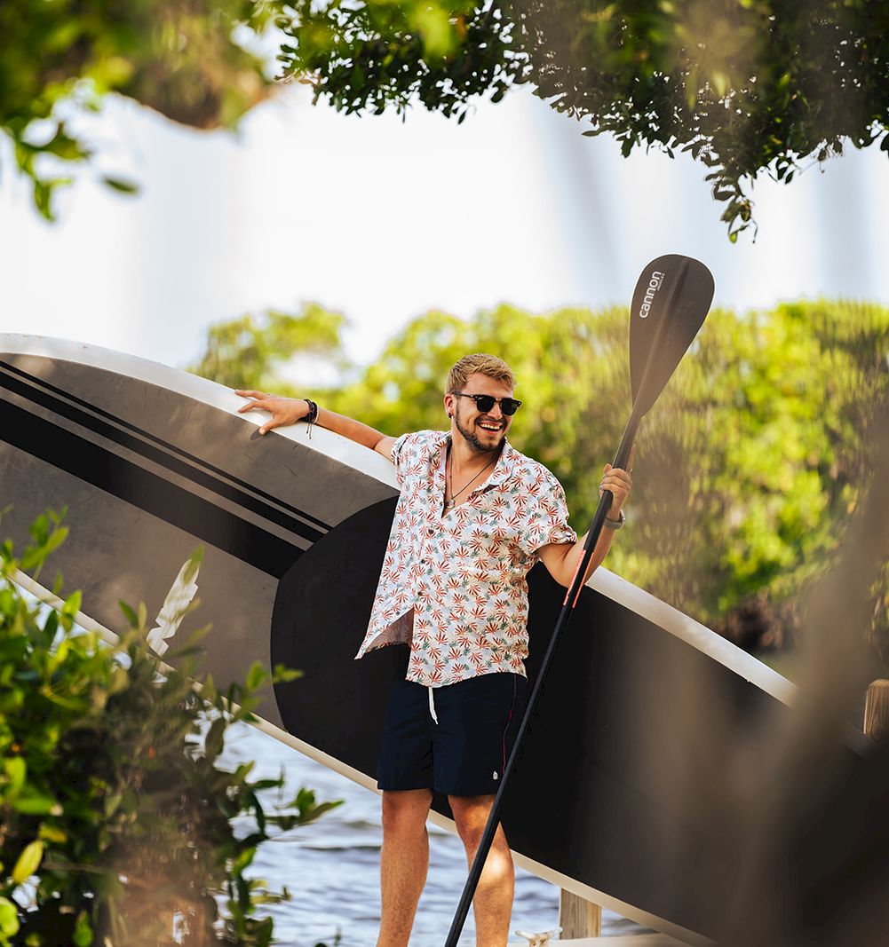 A man carrying a large surfboard and paddle, walking through leafy surroundings on a sunny day.