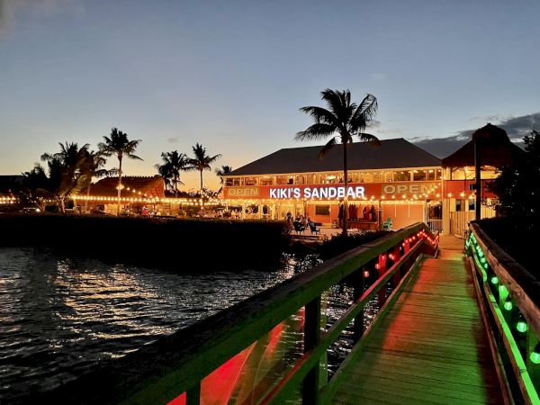 A lively waterfront restaurant on stilts with bright lights, palm trees, and a glowing boardwalk at dusk, inviting diners by the pier.