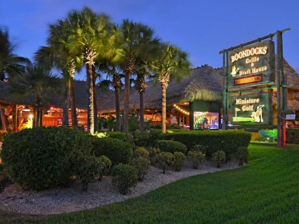 A sunny, tropical restaurant scene with palm trees, manicured bushes, and a lit sign for "Docks" by the water, inviting dining and nightlife.