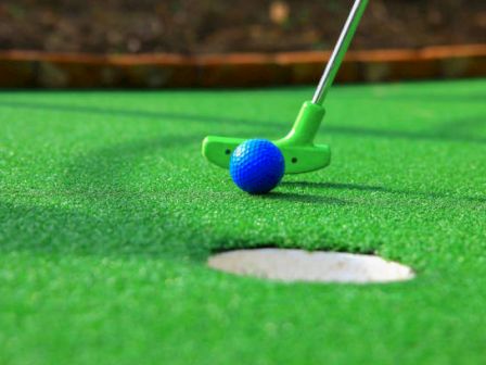 A green mini golf ball rests near the hole, with a putter backing it in on a bright artificial turf green.