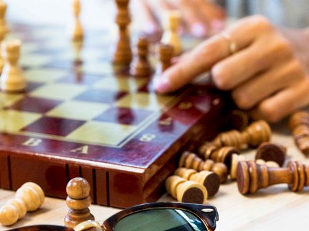 A person plays chess, pieces scattered on a wooden board, sunglasses resting on a light surface, focus on the game&rsquo;s pieces and hands.