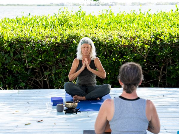 Two people practicing yoga on mats outdoors, facing each other with hands in a prayer pose, on a wooden deck with greenery in the background.