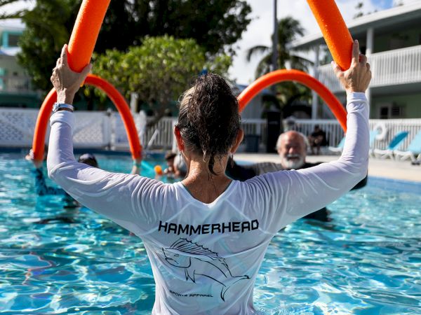 Three people in a pool with orange noodles, the back of a person wearing a "HAMMERCRO" shirt guiding others, sunny day, palm trees and buildings in background.