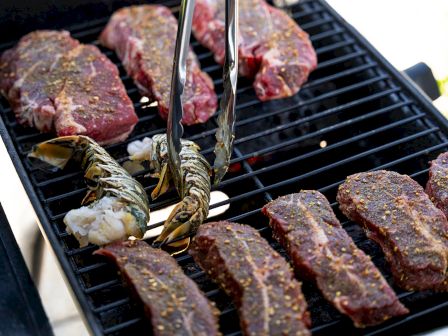 Grilling a variety of meats on a barbecue, including beef steaks and shrimp, with tongs flipping them on the hot grill. End.