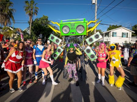 A costumed street parade with a giant green speaker/festival float, dollar-bill wings, and colorful dancers in vibrant outfits marching down the street.