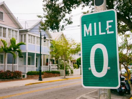 A street scene with pastel houses and palm trees, featuring a large green Mile 0 sign by the sidewalk, marking the start of a route.