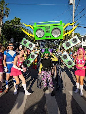 A colorful street parade with people in bright costumes dancing around a large green boombox float adorned with dollar bills, under a sunny sky.
