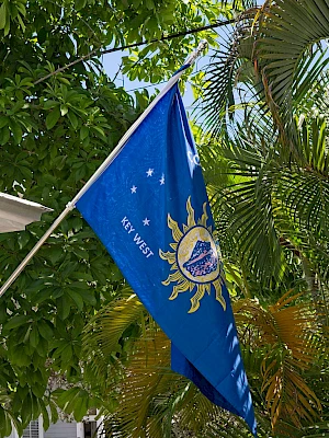 A blue flag with a sun emblem and text flies beside a white porch under leafy green trees, bright daylight outlining tropical scenery.