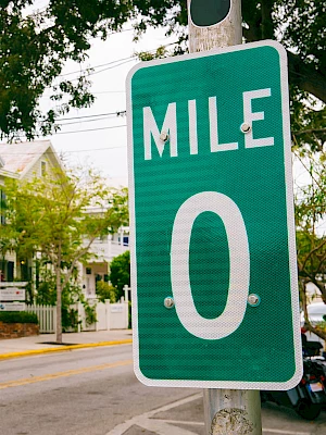 A sunny street with pastel houses and palm trees, a bright green Mile 0 sign on a pole in the foreground.