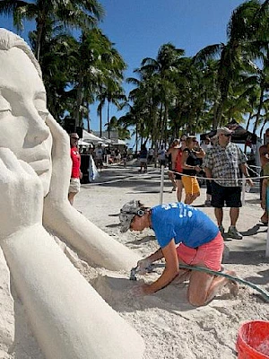 Sand sculpture of a woman with hands under chin, on a beach. A person works on the sculpture; onlookers in swimsuits, palm trees in background.