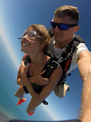 Two people skydiving, smiling and strapped together mid-air, with a bright blue sky and coastline below, selfie-style by the photographer.