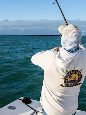 A person wearing a sun-protective white shirt and hood fishes from a boat, holding a rod over calm blue-green sea under a clear sky.