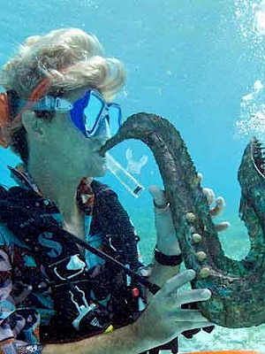 A scuba diver holding a large curved coral or horn-shaped seashell underwater, wearing goggles and a wetsuit, with a blue ocean backdrop.
