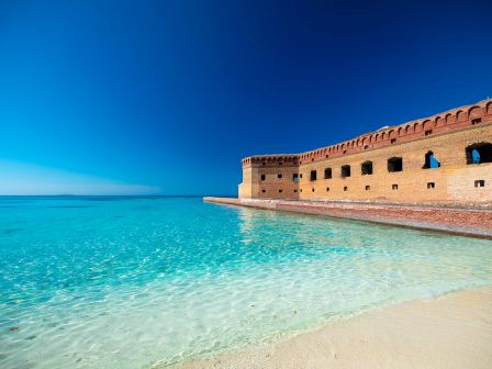 A tropical beach with clear turquoise water, soft sand, and a historic brick fortress stretching along the shore under a bright blue sky.