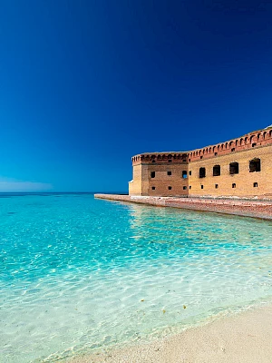 A sunny tropical beach with clear turquoise water lapping a white-sand shore, and an old brick fortress extending along the water’s edge.