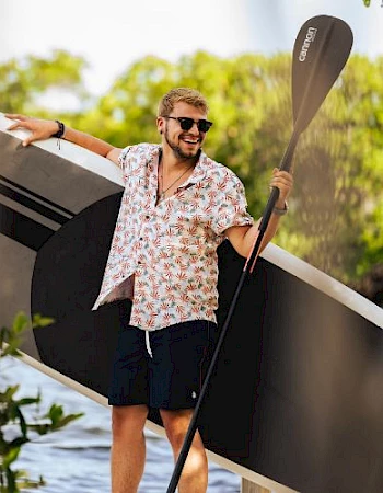 A man in a floral shirt and shorts stands next to a large paddleboard, holding a paddle, outdoors near greenery.