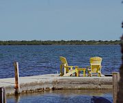 A small seaside dock with bright yellow chairs overlooks calm blue water and distant green shoreline under a clear sky.