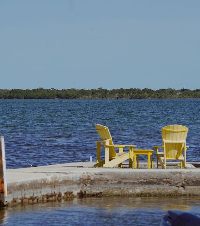 A small seaside dock with bright yellow chairs overlooks calm blue water and distant green shoreline under a clear sky.
