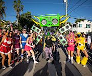 A colorful street parade with dancers and a giant green boombox robot float amid sunny palms and cheering crowds.