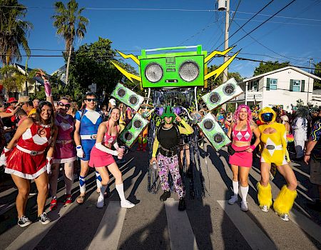 A colorful street parade with dancers and a giant green boombox robot float amid sunny palms and cheering crowds.